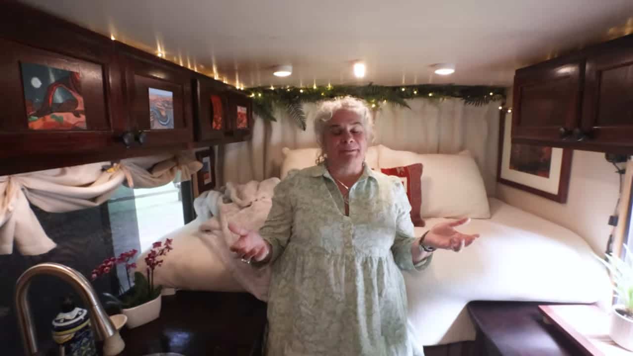Eva standing by the cozy white bed with fairy lights and greenery garland in Box Truck Betti