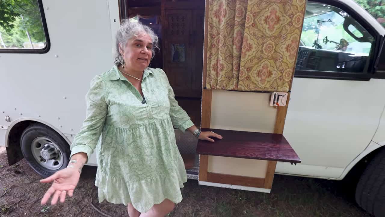 Eva standing at the open side door of her white box truck tiny home with a wooden fold-out table
