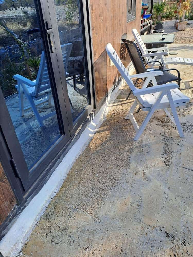 Masonry enclosing the gap between the expandable prefab home and its concrete base, beside glass doors and white patio chairs