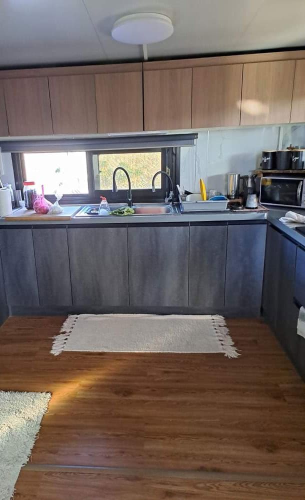 Kitchen in the expandable prefab home in Cyprus with wood-look upper cabinets, grey lower cabinets, a stainless sink under a wide window and a wood-look floor