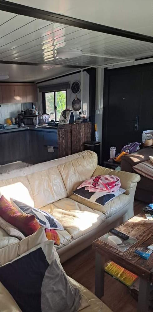 Open living space in the expandable prefab home in Cyprus, a cream leather sofa and wooden coffee table in front, with the kitchen and a window beyond