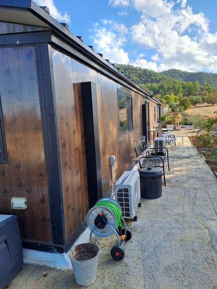 Side of the expandable prefab home in Klirou, Cyprus, with wood-look cladding, an air-conditioning unit and hose reel along a concrete path, and wooded hills behind