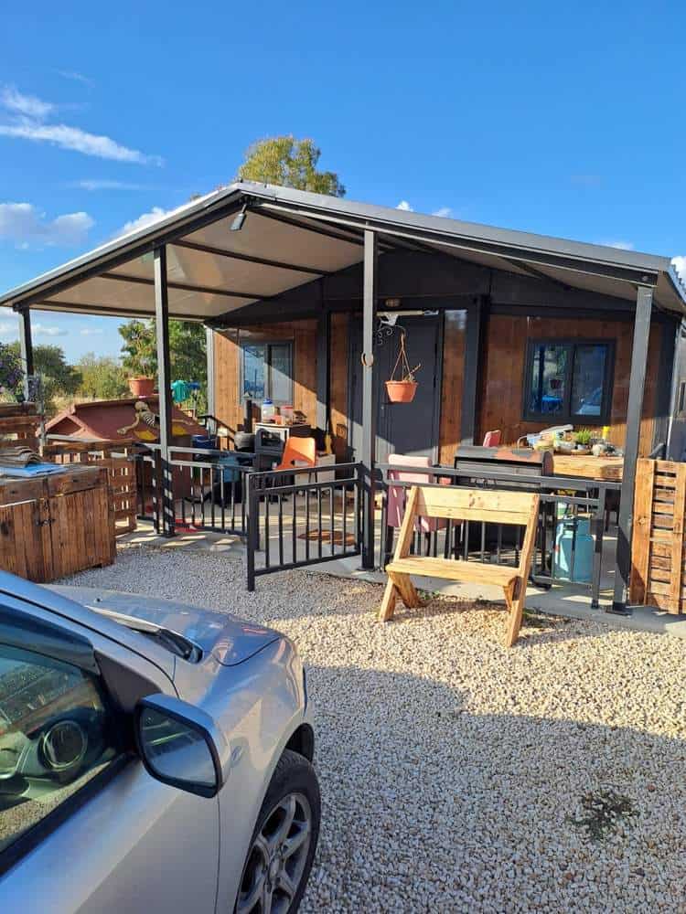 Expandable prefab family home in Klirou, Cyprus, with dark wood-look cladding under an added roof and covered veranda, a pallet-wood bench on the gravel in front