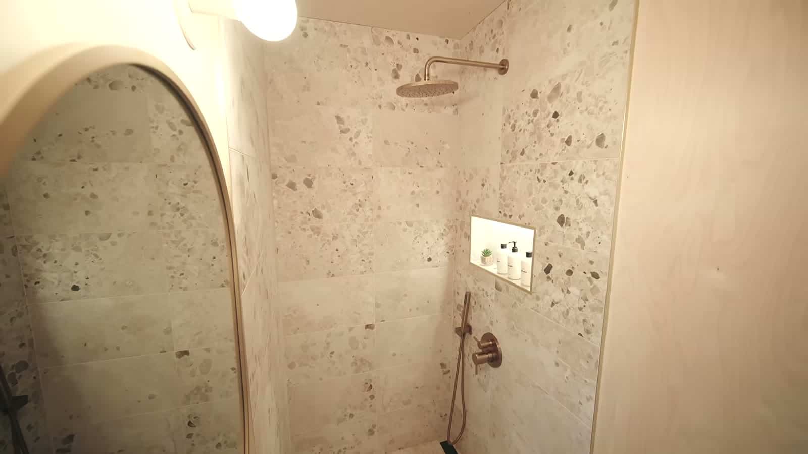 Walk-in shower in the B.Drop.24f with terrazzo-look tile, an arched opening, brass rain head and a recessed toiletry niche