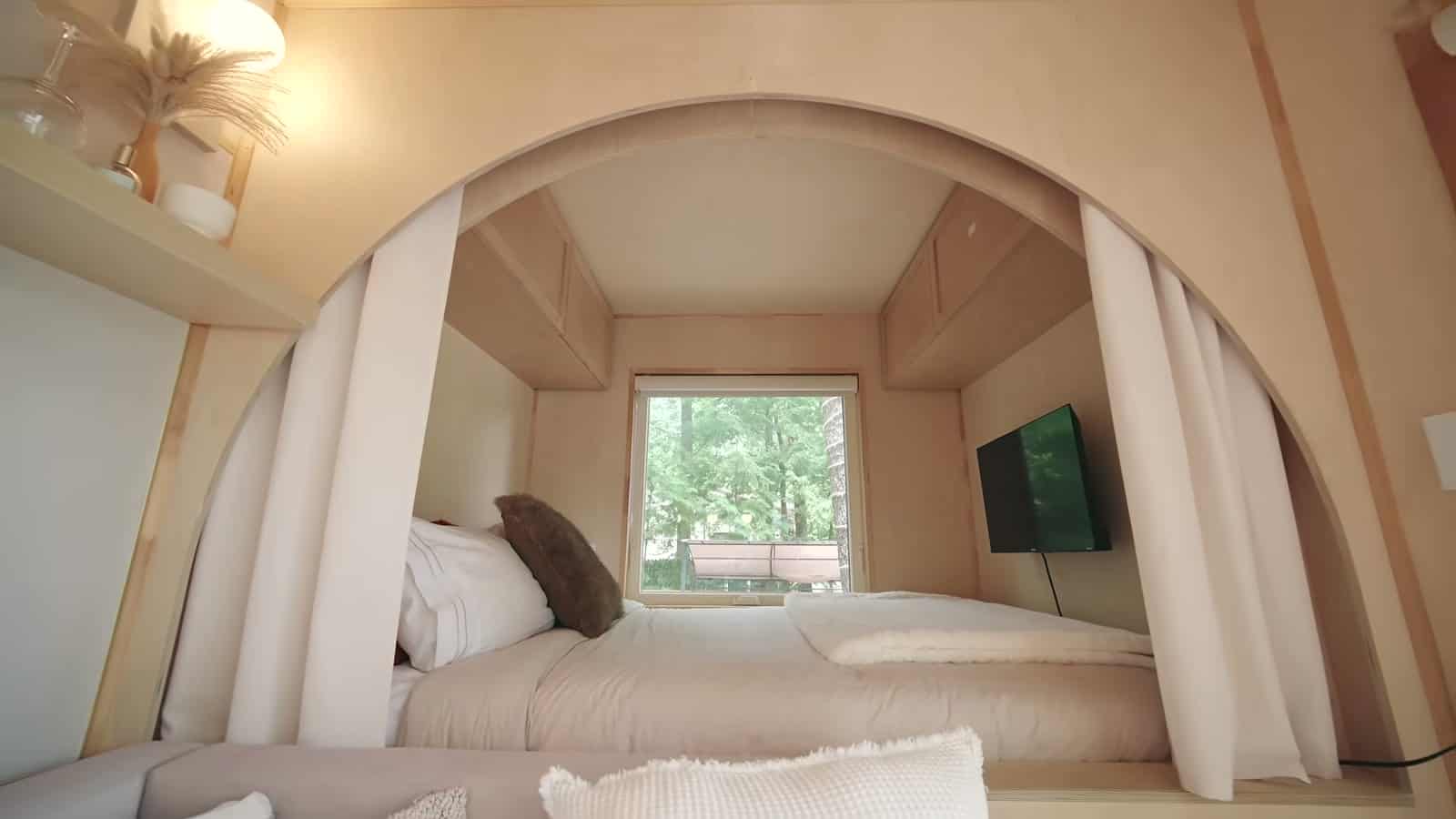 Raised queen bed in the arched alcove of the B.Drop.24f, curtains pulled back, forest-facing window and a wall-mounted TV