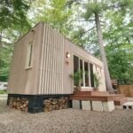 Be Tiny B.Drop.24f tiny house in a wooded campground, vertical cedar cladding under an angled shed roof, firewood stacked beneath the cedar deck