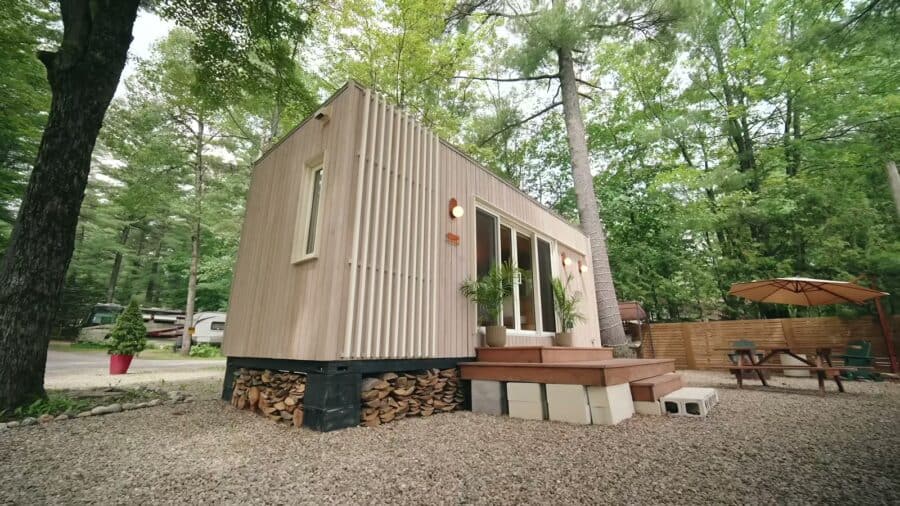 Be Tiny B.Drop.24f tiny house in a wooded campground, vertical cedar cladding under an angled shed roof, firewood stacked beneath the cedar deck