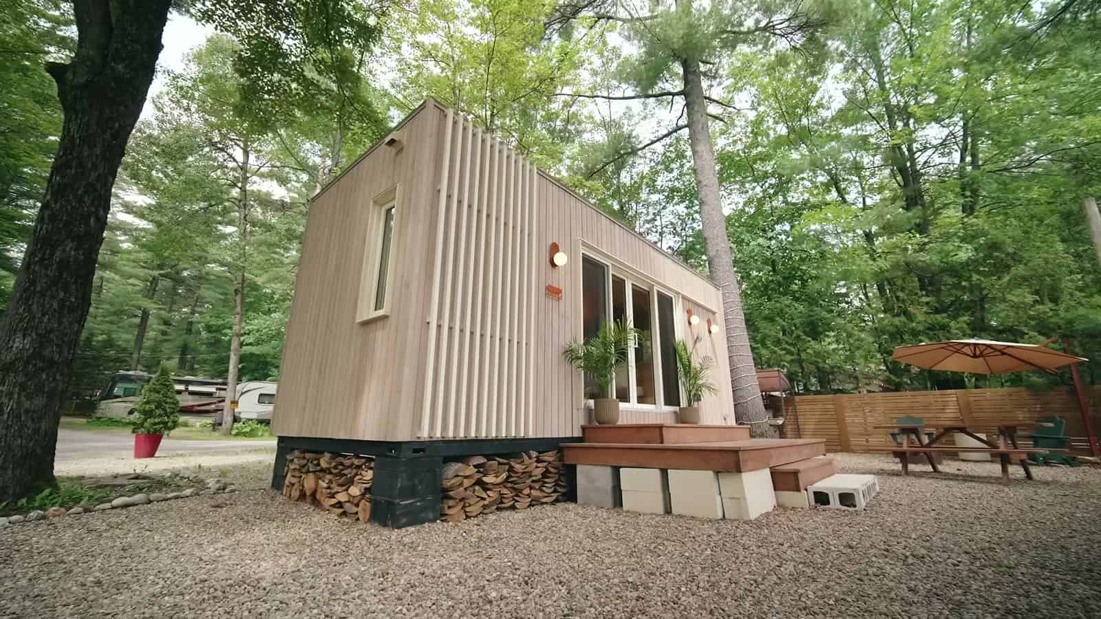 Be Tiny B.Drop.24f tiny house in a wooded campground, vertical cedar cladding under an angled shed roof, firewood stacked beneath the cedar deck