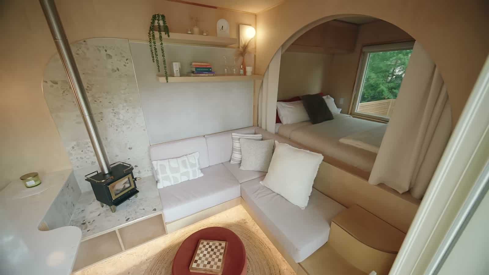 Small black wood stove on a terrazzo hearth wall beside the L-shaped sofa, with the arched opening to the raised bed behind