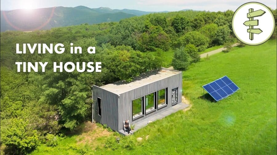 Gray plank off-grid studio on pillars with a wooden deck and solar array in a meadow, forest and mountains beyond
