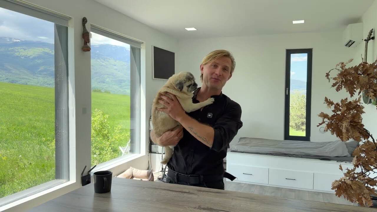 Ivan holding his dog in the bright studio, with the platform bed and big windows to the meadow and mountains behind him