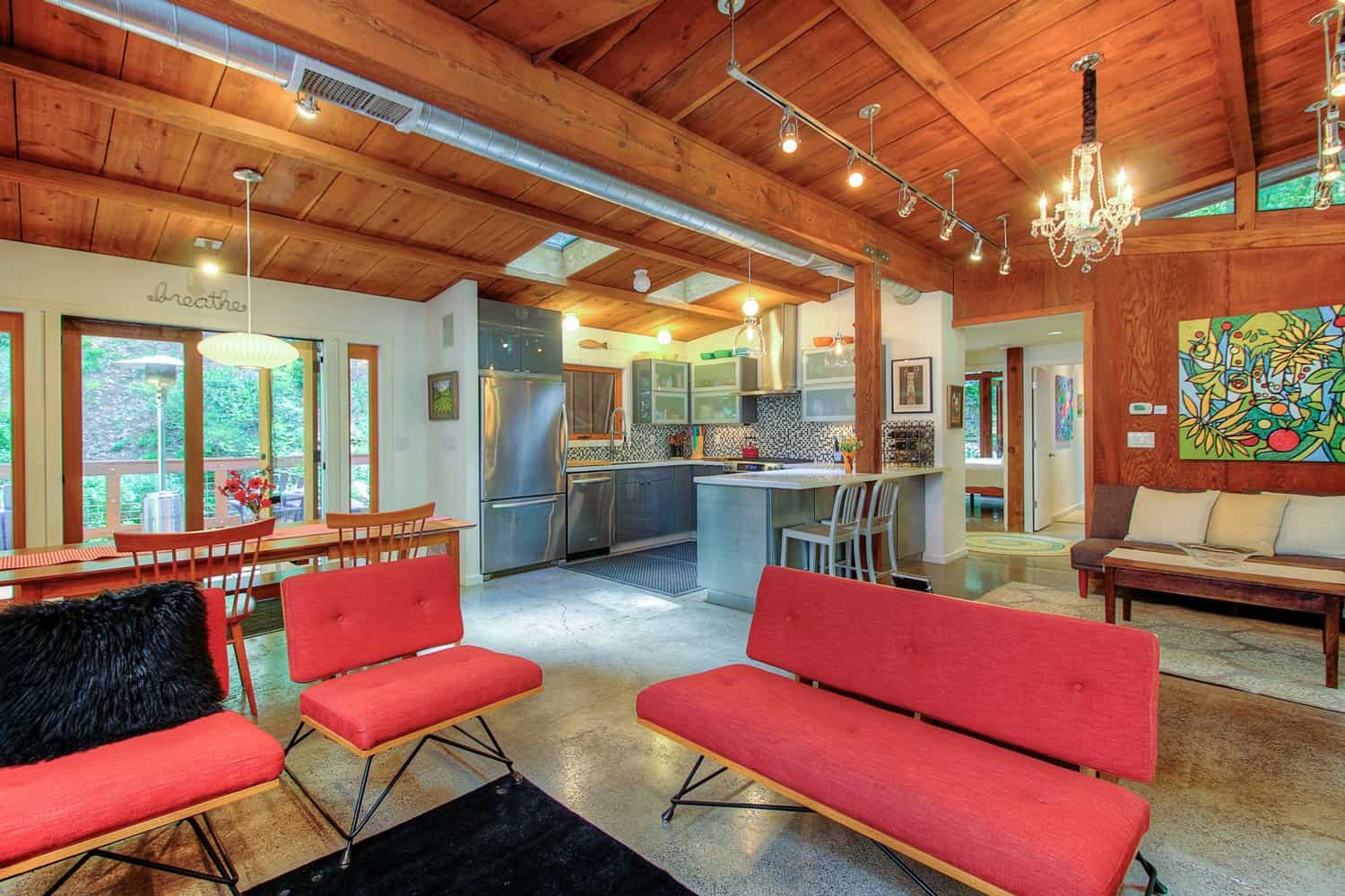 Open living area with red mid-century sofas, wood ceiling, skylights, and windows facing the trees