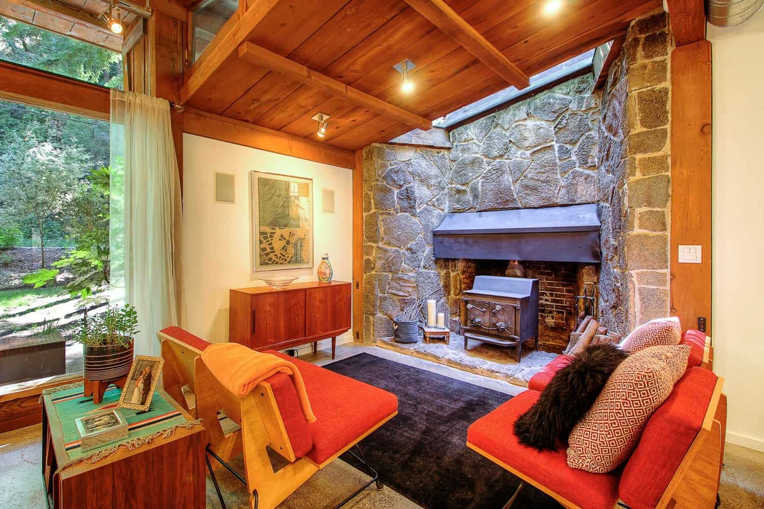 Living room with a large stone fireplace and wood stove, wood-beamed ceiling, and colorful mid-century furniture