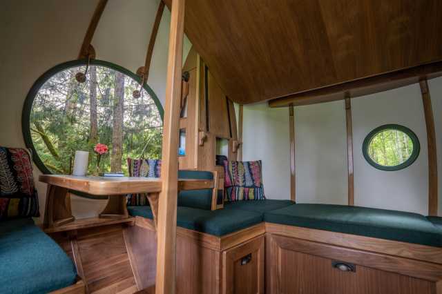Wood-ribbed Luna interior: wrap-around green benches, colorful pillows, circular forest windows, flat wood panel of the raised hydraulic bed