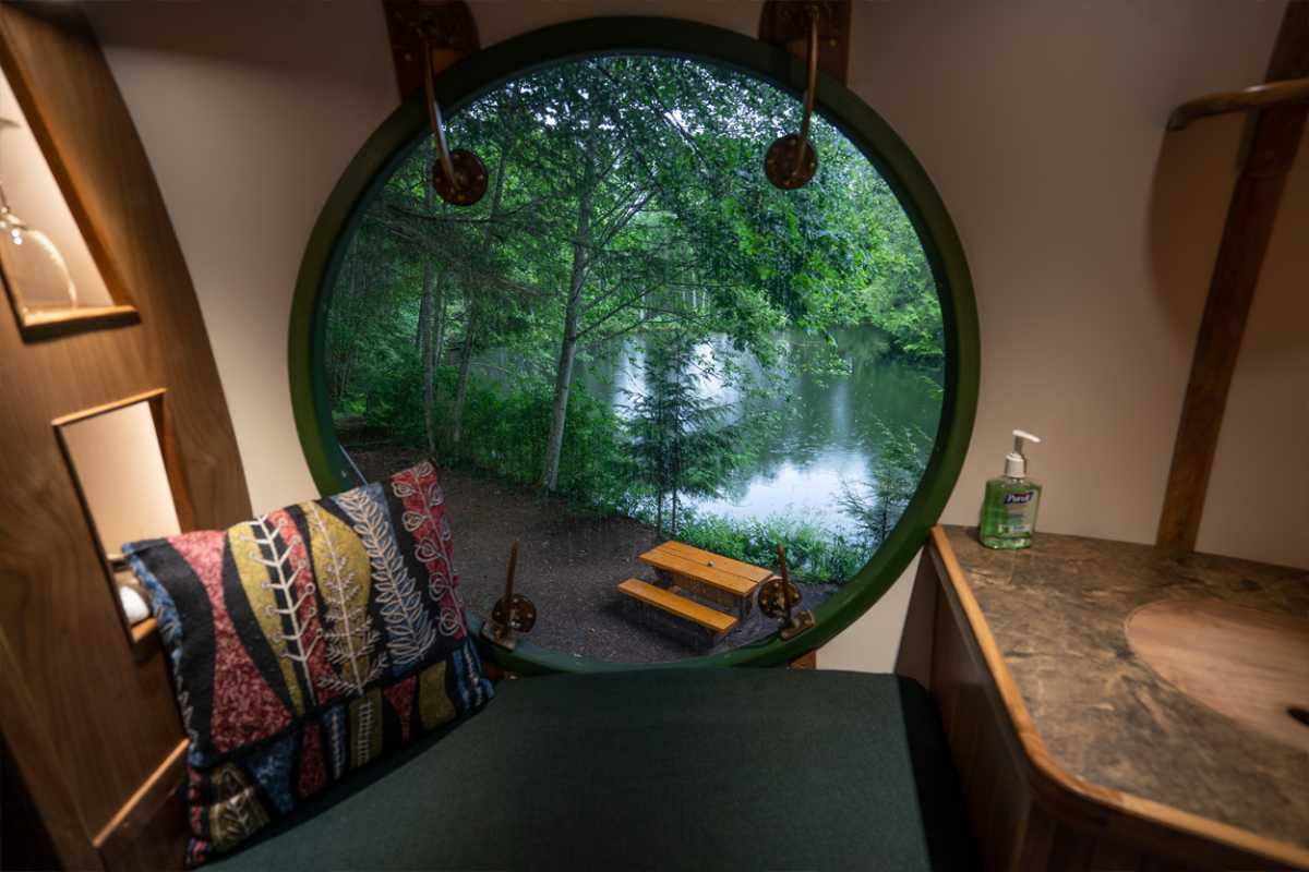 Luna interior: circular window over a green cushioned bench, galley counter with a round wood sink, forest and a picnic table outside