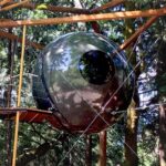 Dark glossy Free Spirit Spheres Luna hanging among tall trees: one circular porthole, tension cables to a wood frame, a small plank landing to the left