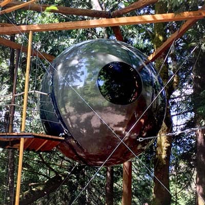 Dark glossy Free Spirit Spheres Luna hanging among tall trees: one circular porthole, tension cables to a wood frame, a small plank landing to the left