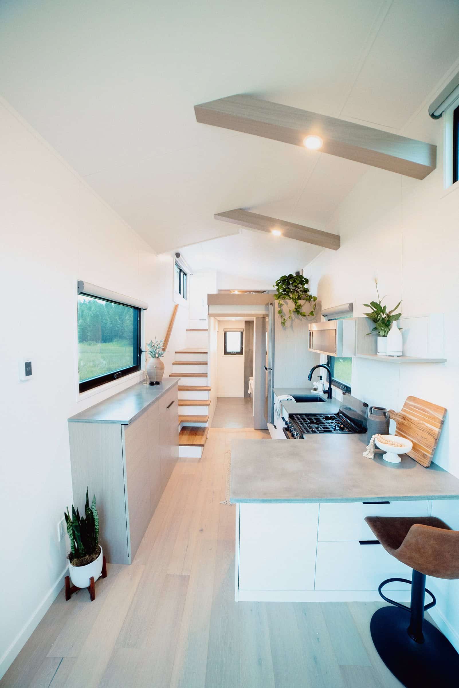Halcyon 01 kitchen: concrete-style counters, white cabinets, storage stairs to the loft, wood beam ceiling, forest window