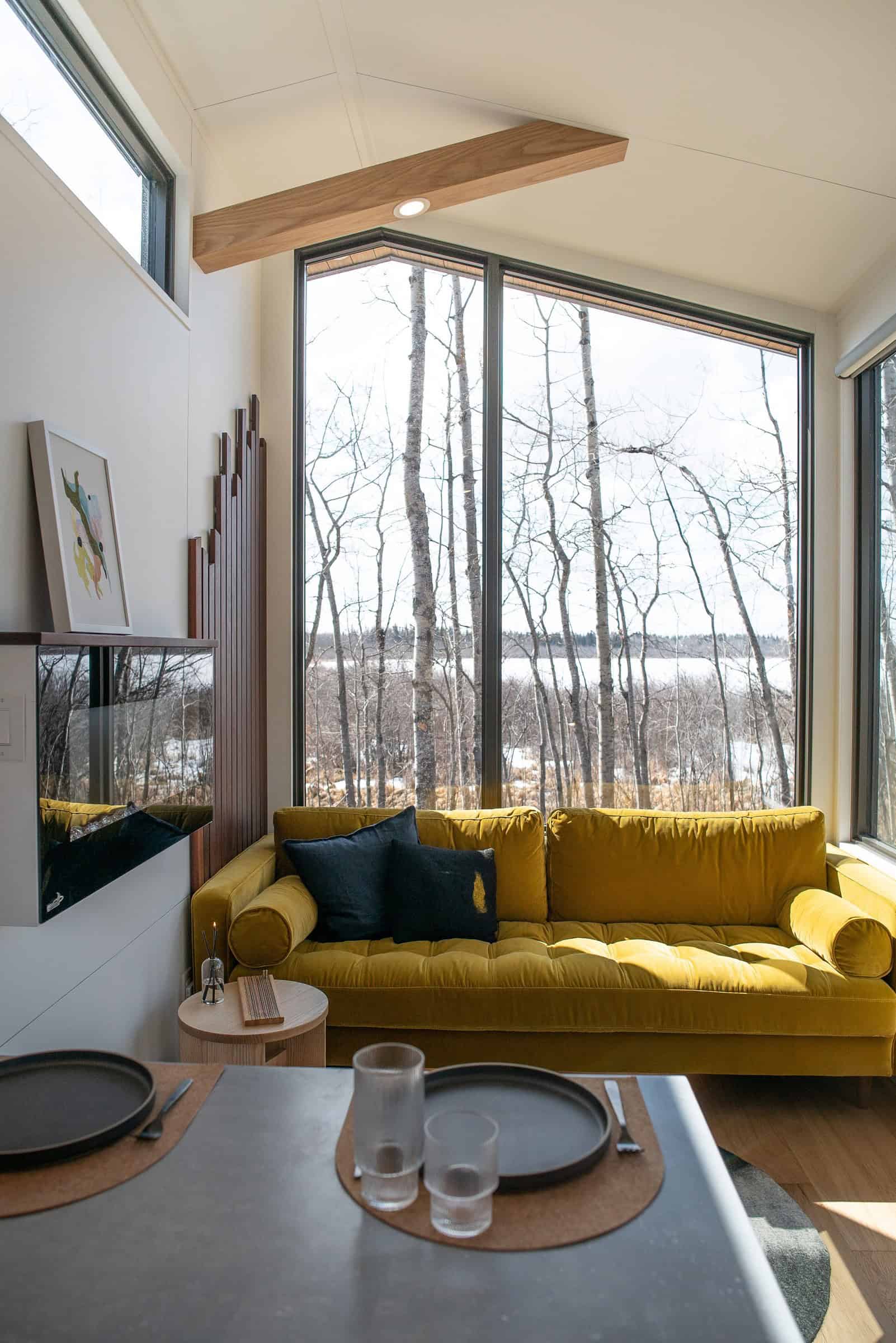 Halcyon 01 living: mustard sofa, floor-to-ceiling winter windows, white vaulted ceiling with a wood beam, light wood floor