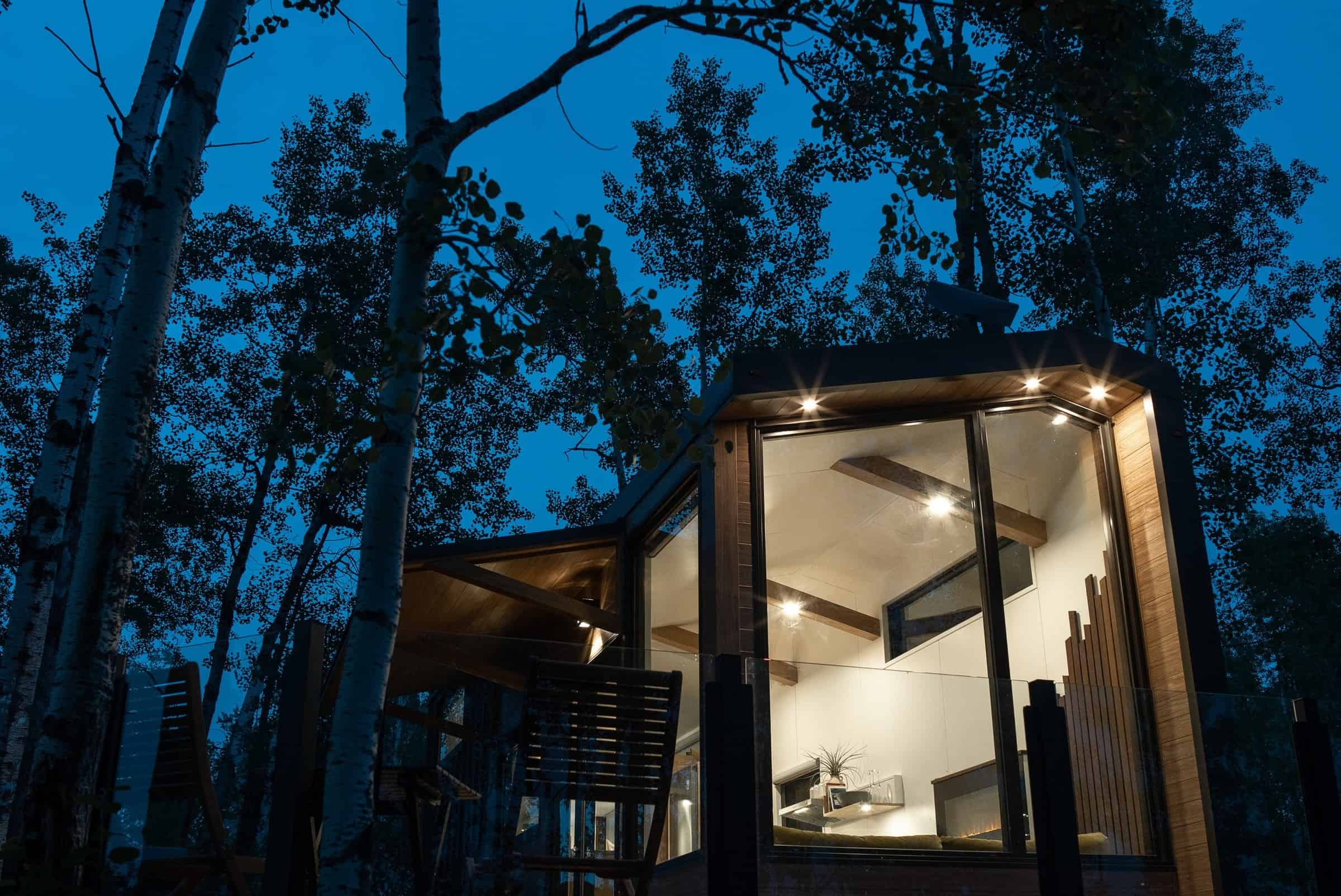 Halcyon 01 at twilight among birch: floor-to-ceiling glass glowing, soffit lights on the overhang, glass-rail deck
