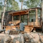 Wider Halcyon 01 exterior: light wood and dark metal siding, glass-rail deck, gravel patio with a fire pit and Adirondack chairs, birch trees