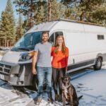 Couple with dog in front of a DIY van conversion in snowy forest setting.