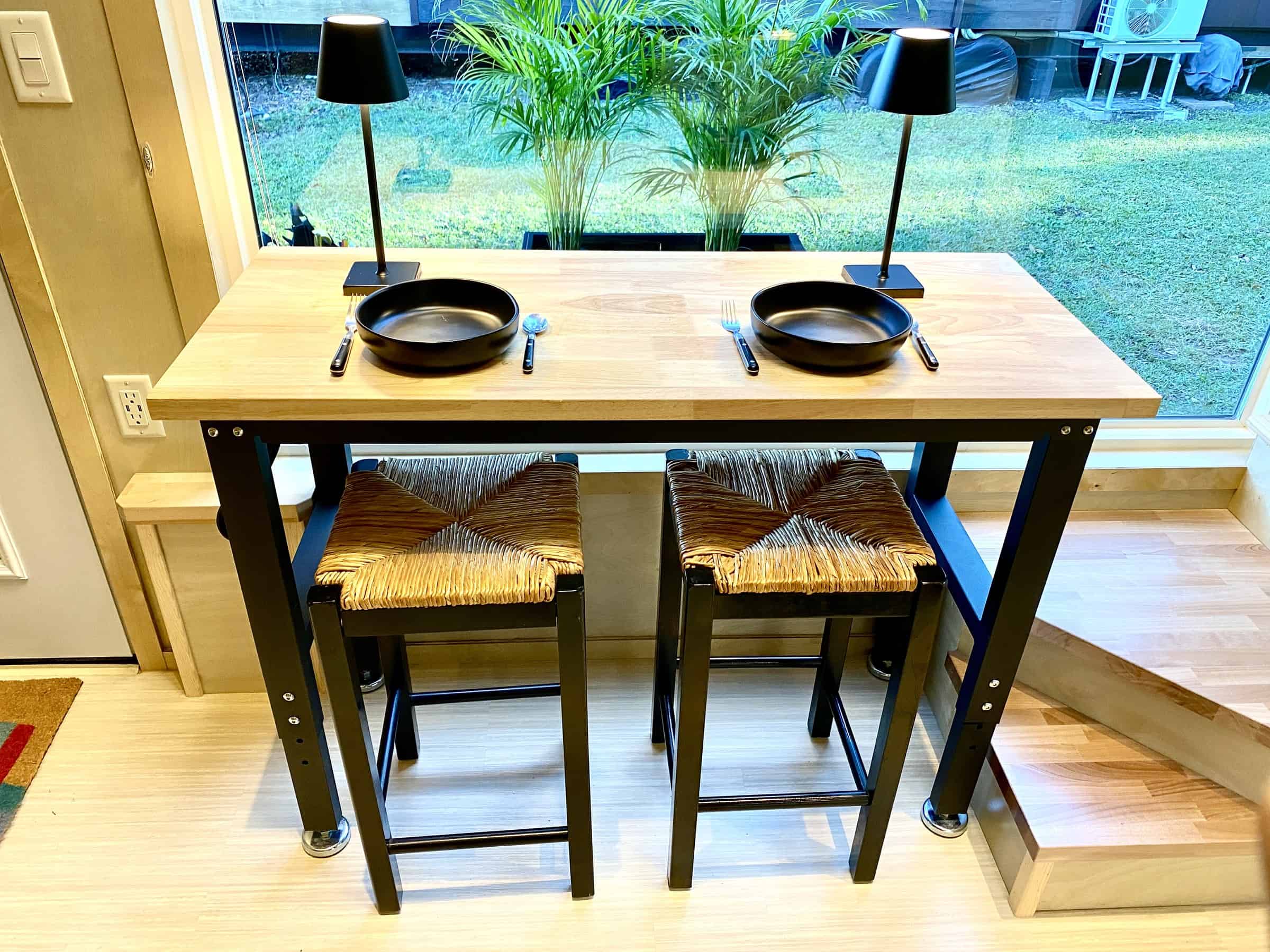 Counter-height wood dining table with two woven stools set for two in front of a large window, birch walls and light plank floors