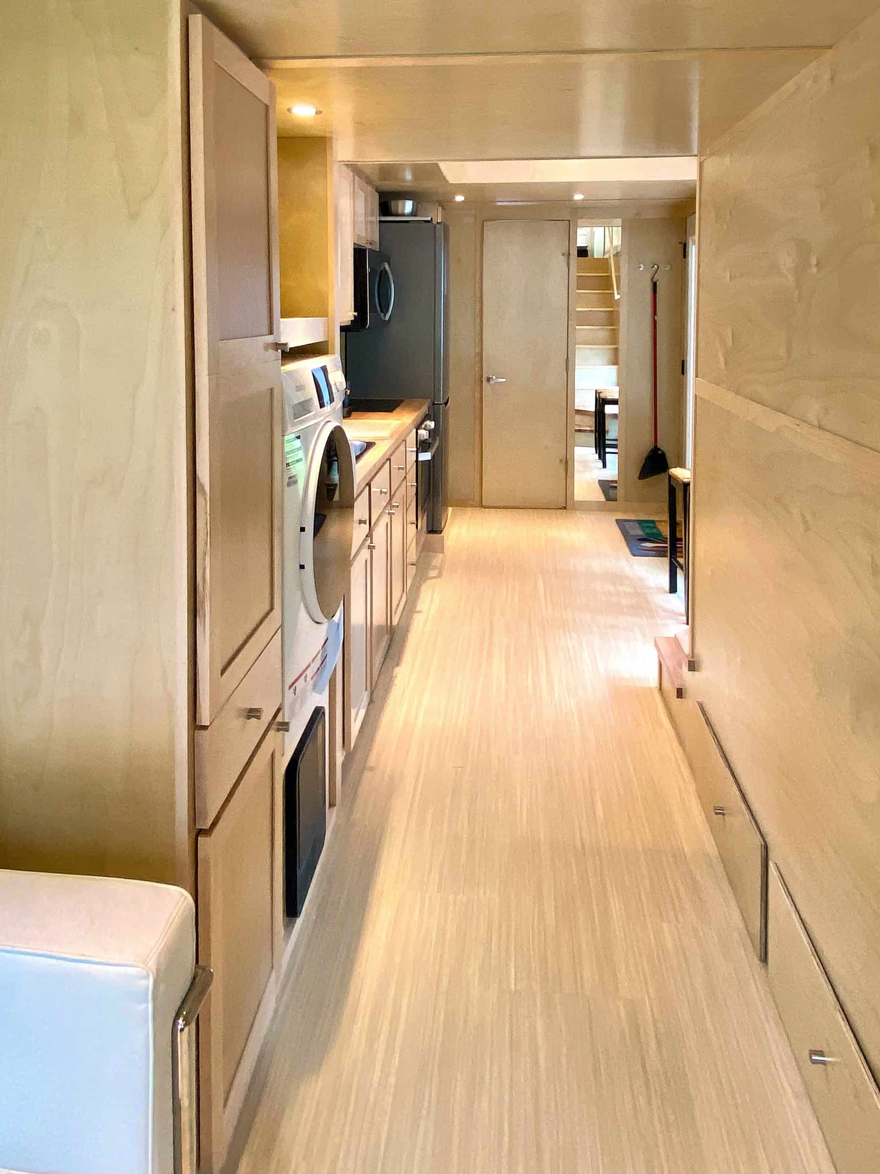 Hallway of the eONE with extra-tall birch cabinets, a white washer-dryer combo, galley kitchen and tall fridge on the left, low storage drawers on the right, stairs and living light at the far end