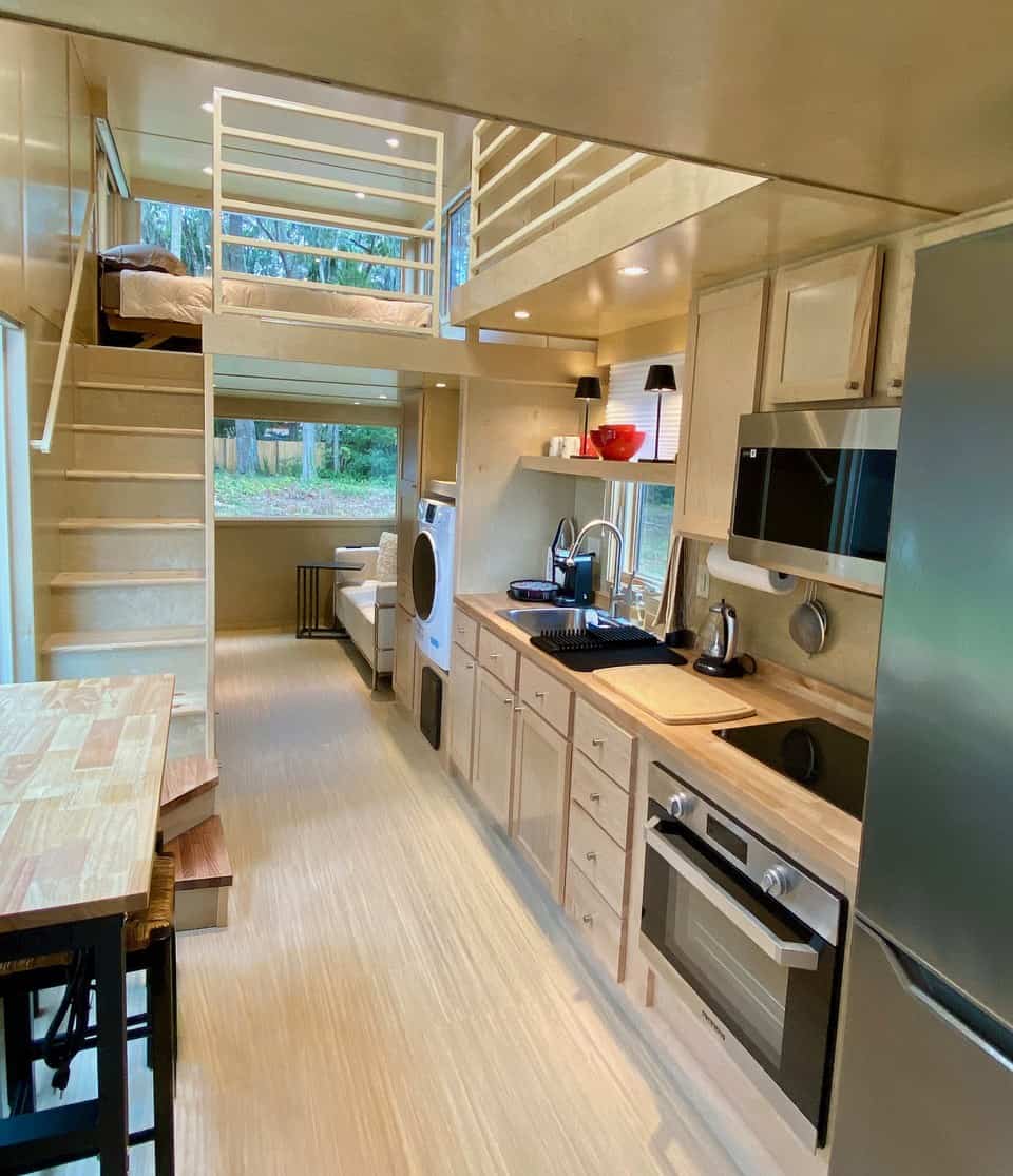 Looking down the birch eONE: butcher-block kitchen with induction cooktop, oven, microwave and tall fridge, washer-dryer combo beyond, living window at the end, and a loft with a white railing above