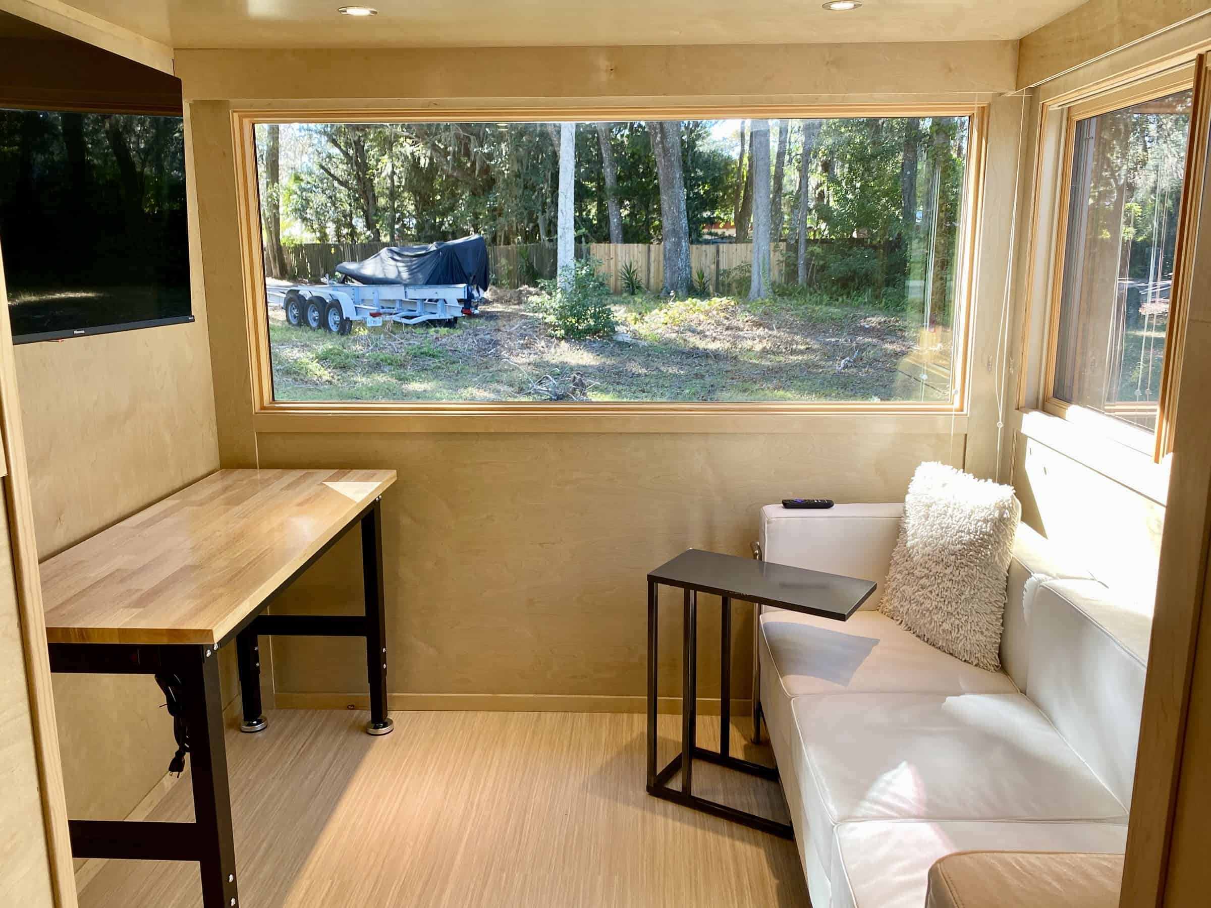 Birch-paneled living end of the eONE with a butcher-block desk, a wall-mounted 43-inch smart TV, a white loveseat, and a large picture window looking onto grass and trees