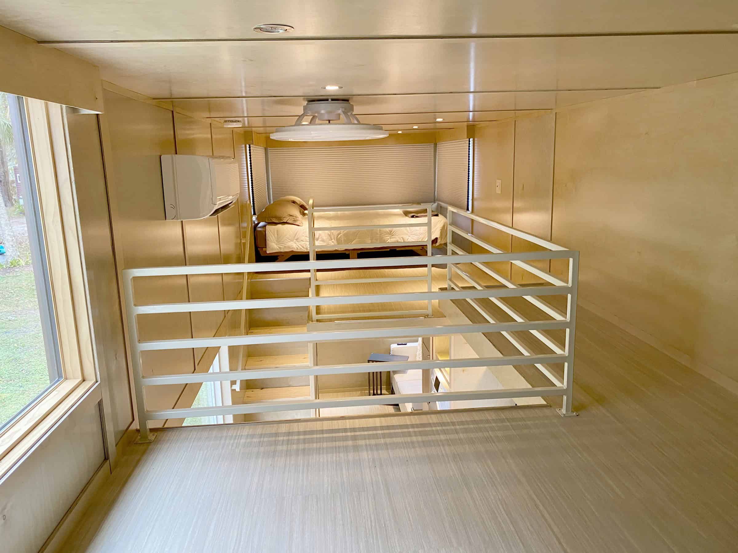 Looking across the eONE walkway to the sleeping loft, a bed with tan bedding and white pleated shades on three large windows, mini-split and living room visible below