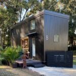 Dark-sided ESCAPE eONE tiny house on a New Smyrna Beach lot, glass entry door under a small metal awning, kitchen window, loft glass, wooden deck with palms, and site number 8