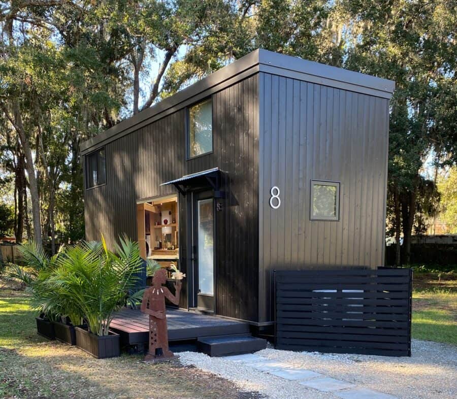 Dark-sided ESCAPE eONE tiny house on a New Smyrna Beach lot, glass entry door under a small metal awning, kitchen window, loft glass, wooden deck with palms, and site number 8