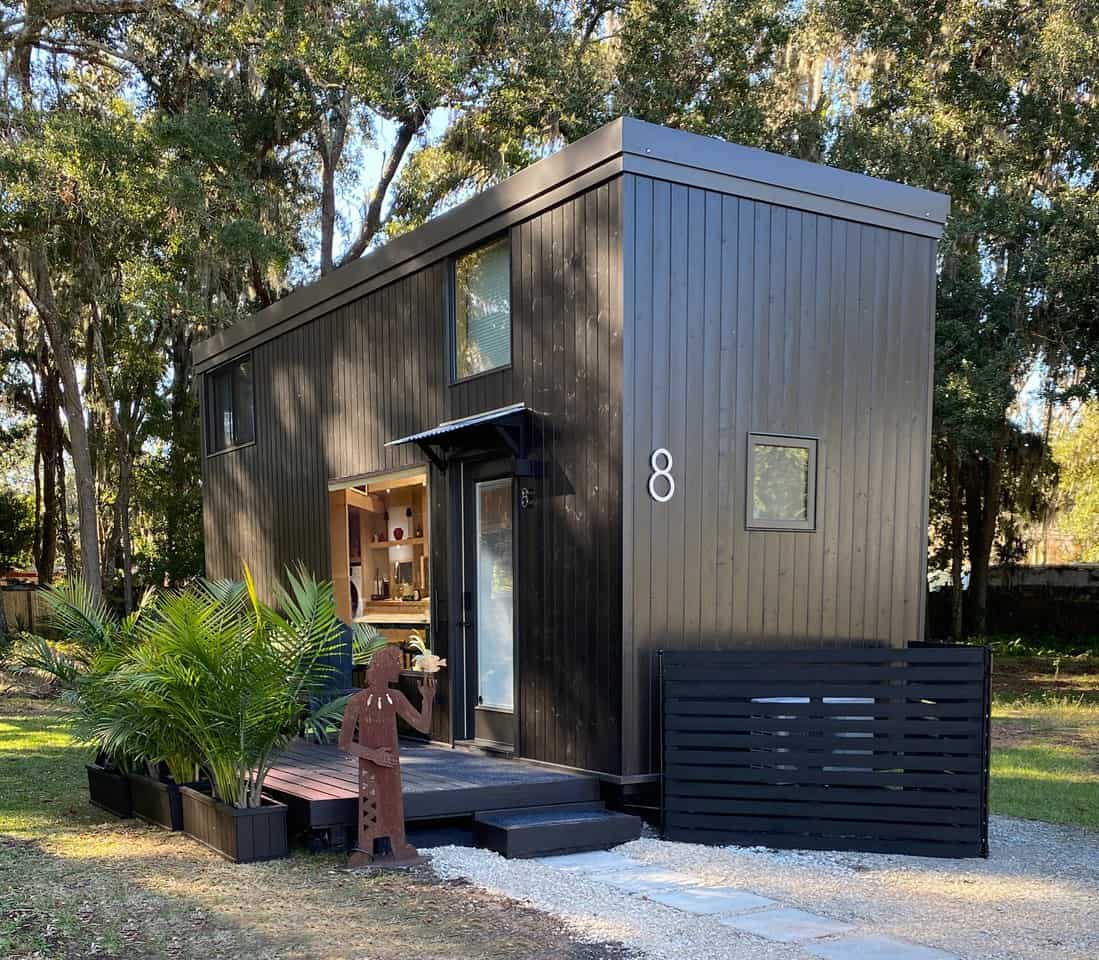 Dark-sided ESCAPE eONE tiny house on a New Smyrna Beach lot, glass entry door under a small metal awning, kitchen window, loft glass, wooden deck with palms, and site number 8