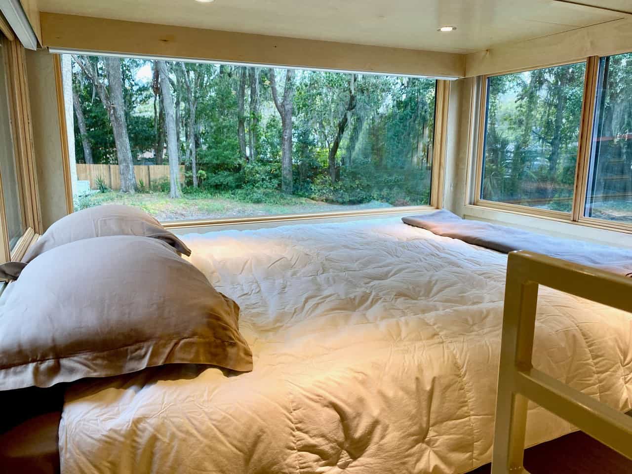 Queen bed in the eONE loft surrounded by large windows with white blockout shades, birch walls and recessed ceiling lights, trees and Spanish moss outside