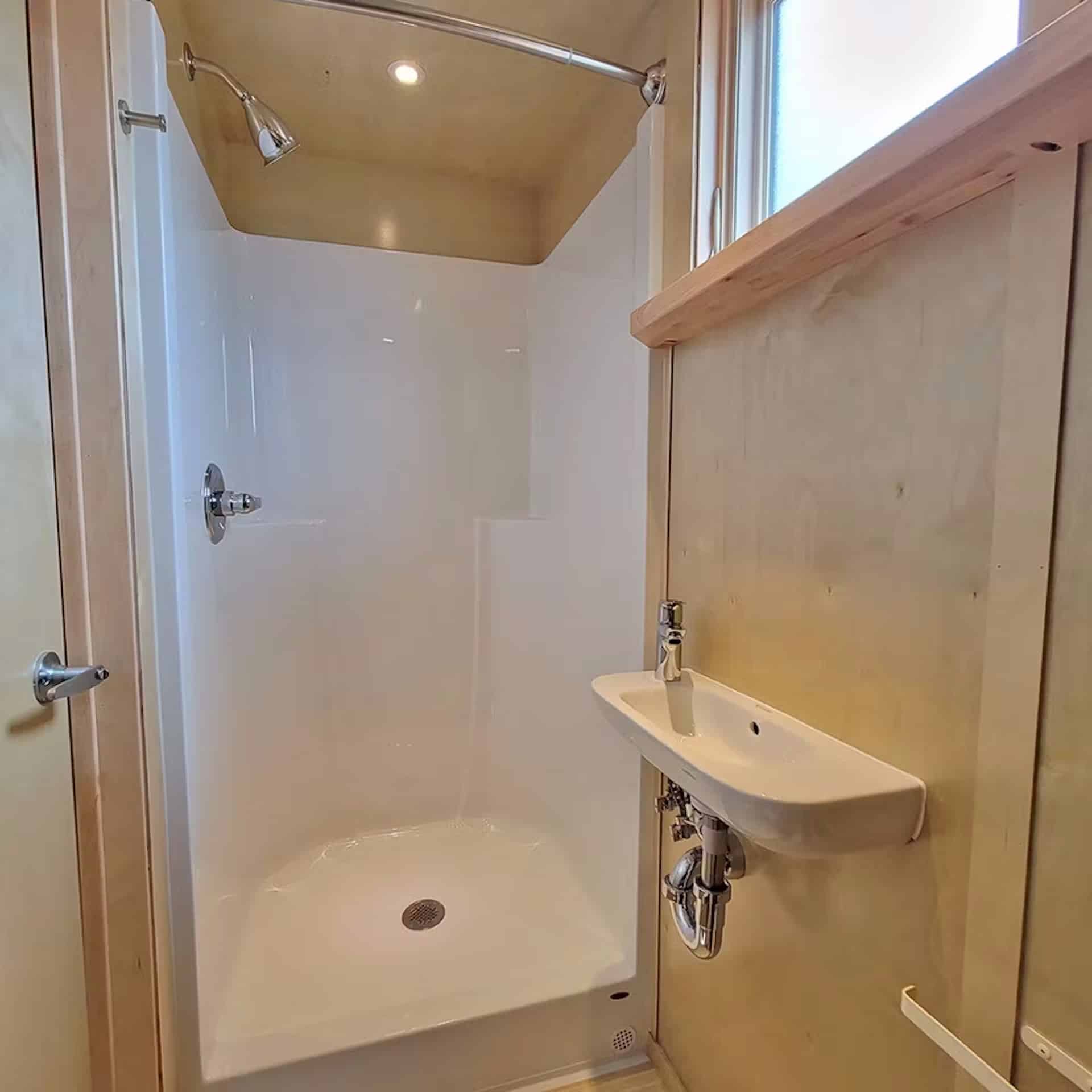 Full-size white shower stall and a wall-mounted sink in a birch-paneled bathroom with a window over a wood shelf