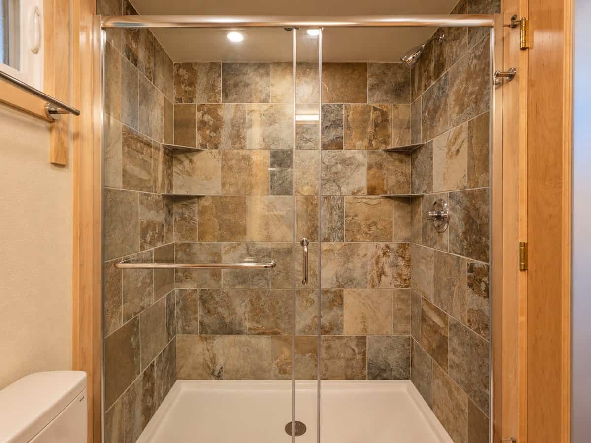 Custom tiled shower with natural stone-look tile, corner shelves and a glass enclosure in the GLASS HOUSE Lot 25