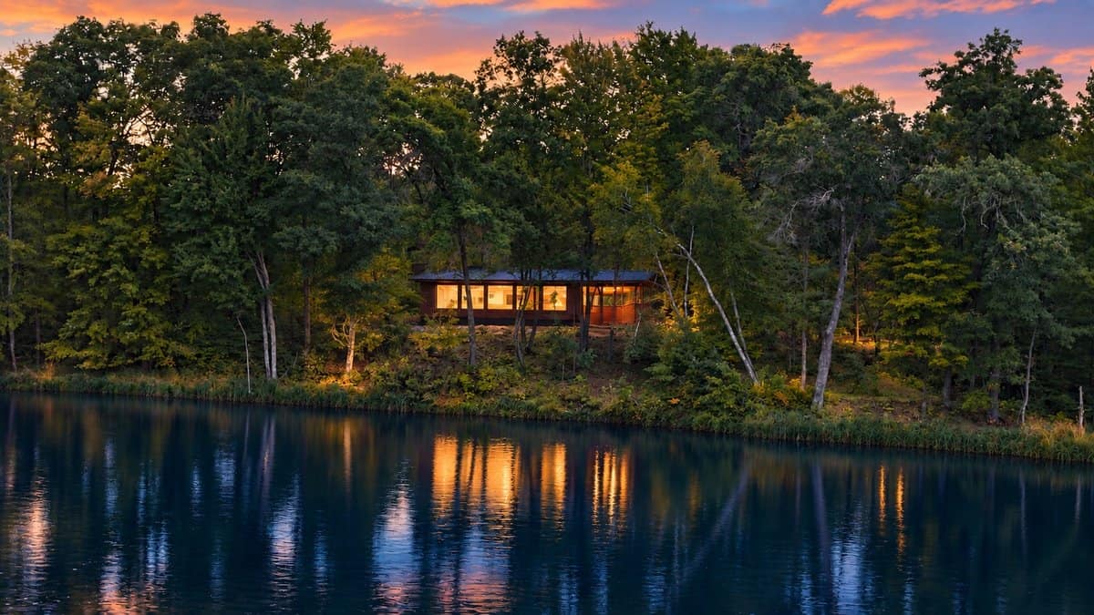 The GLASS HOUSE glowing at dusk on a wooded rise above Lost Lake, seen from across the water at Canoe Bay Village Lot 25