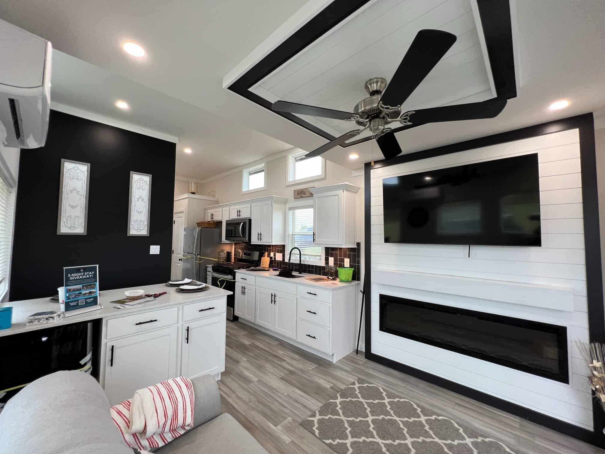 Bayview 300 living: white shiplap fireplace/TV wall, tray ceiling with black fan, grey sofa, open kitchen beyond