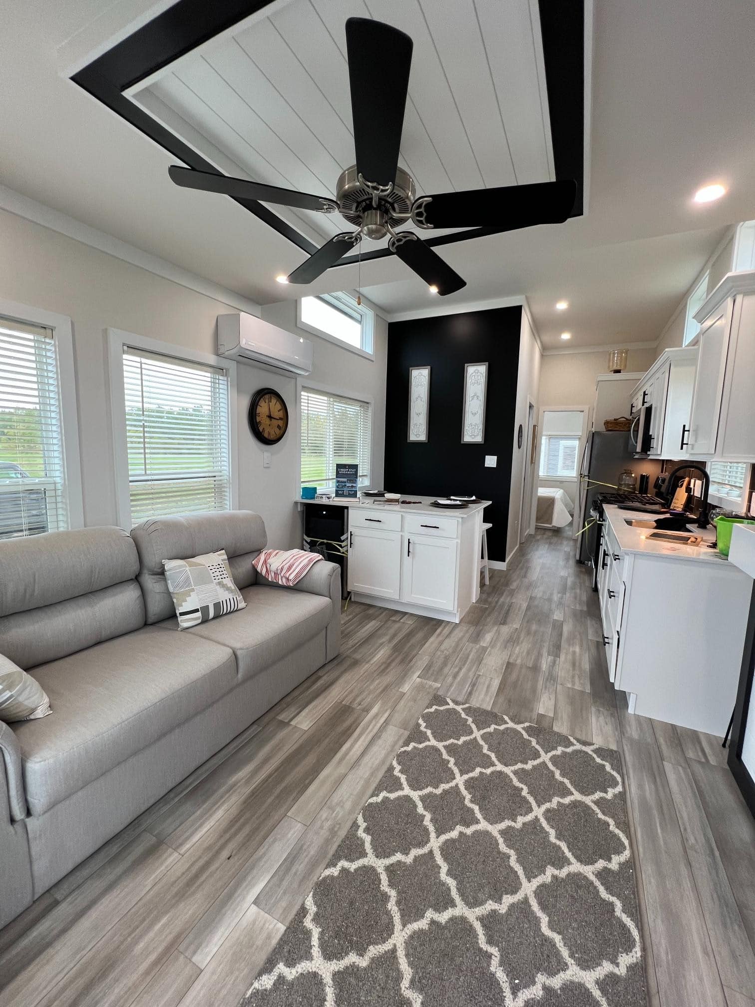 Living-to-kitchen run in Bayview 300: grey sofa, white island with wine fridge, black accent wall, bedroom doorway ahead