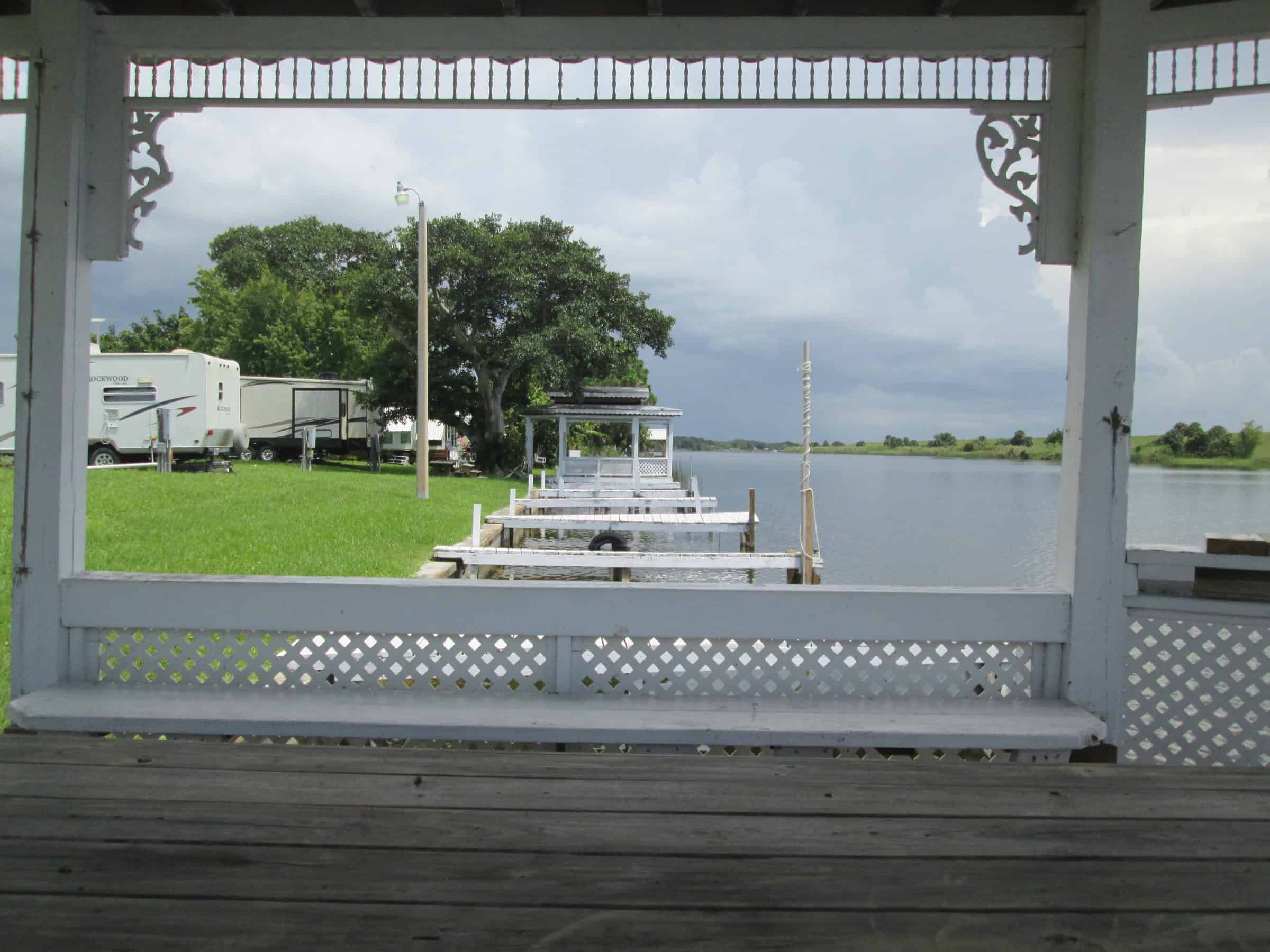 View from inside a white gazebo toward a row of boat docks on the canal, with RVs parked on the lawn at left