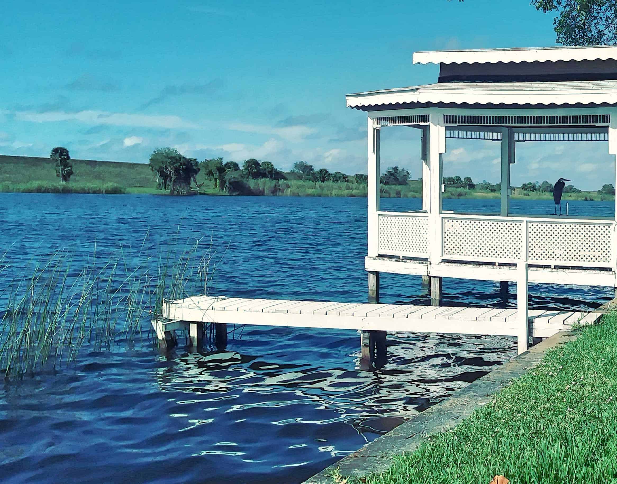 White gazebo and wooden dock over blue water with a heron standing on the gazebo rail