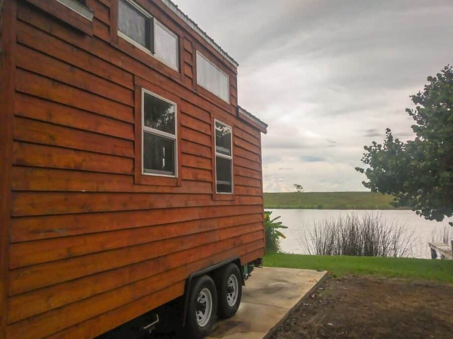 Side of a cedar-sided tiny house on wheels parked on a concrete pad with the canal and levee in the background