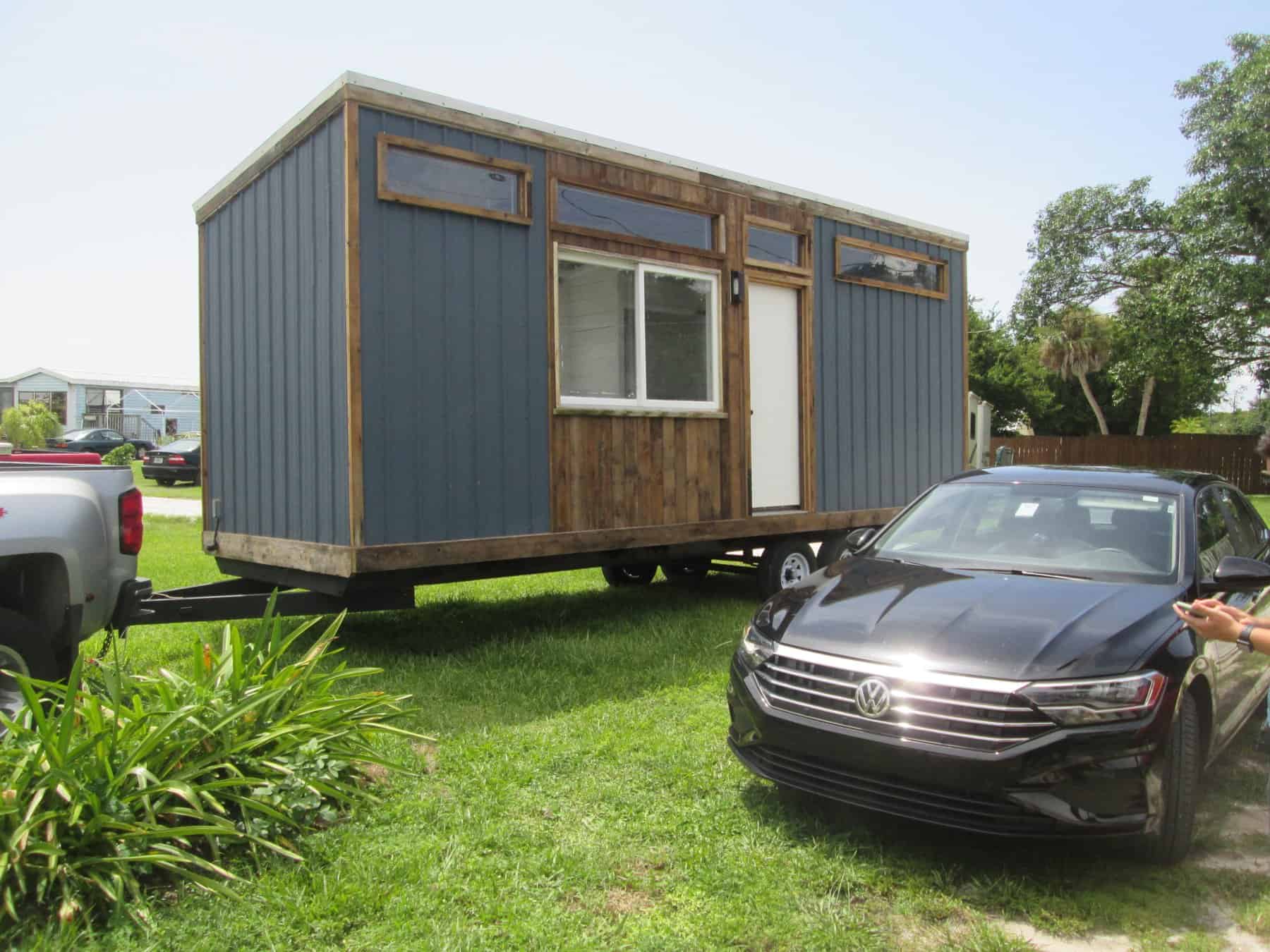 Blue and wood-sided tiny house on wheels parked on a grassy site with a car beside it