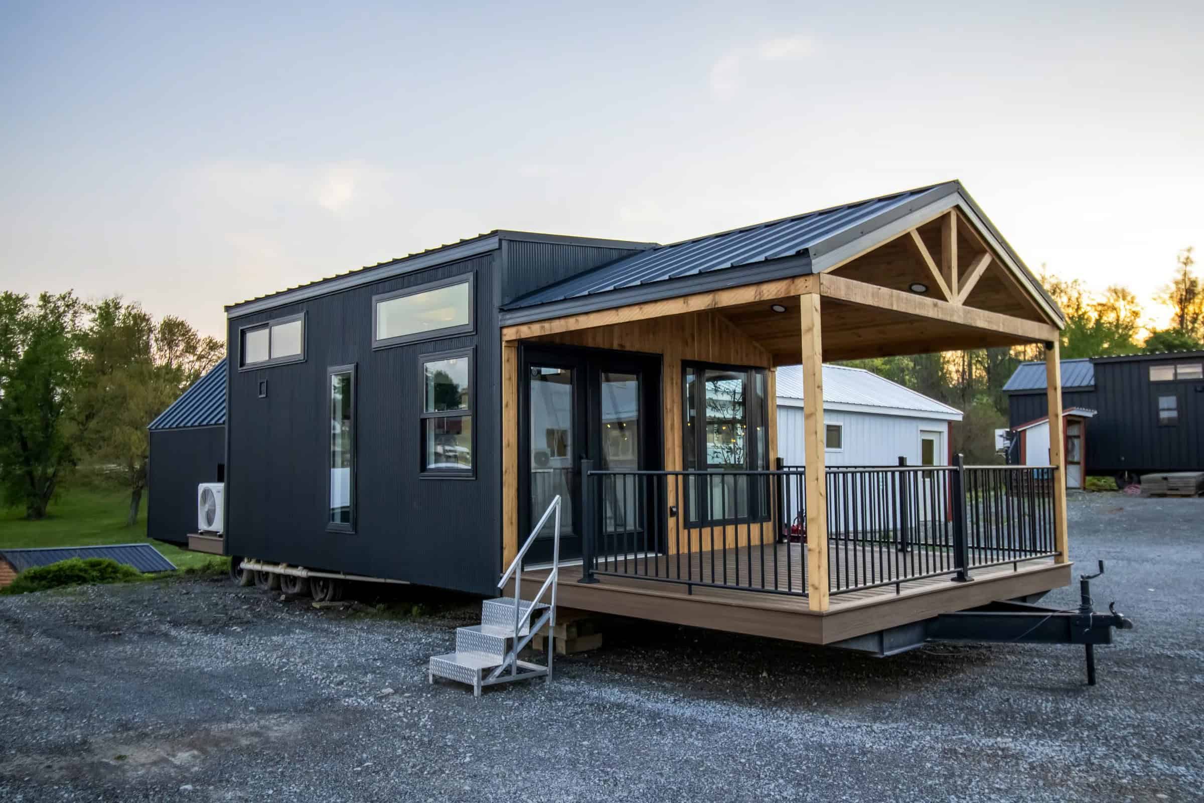 Front three-quarter of Great Lakes A-Frame Luxe: timber porch, black metal roof, trailer hitch on gravel lot