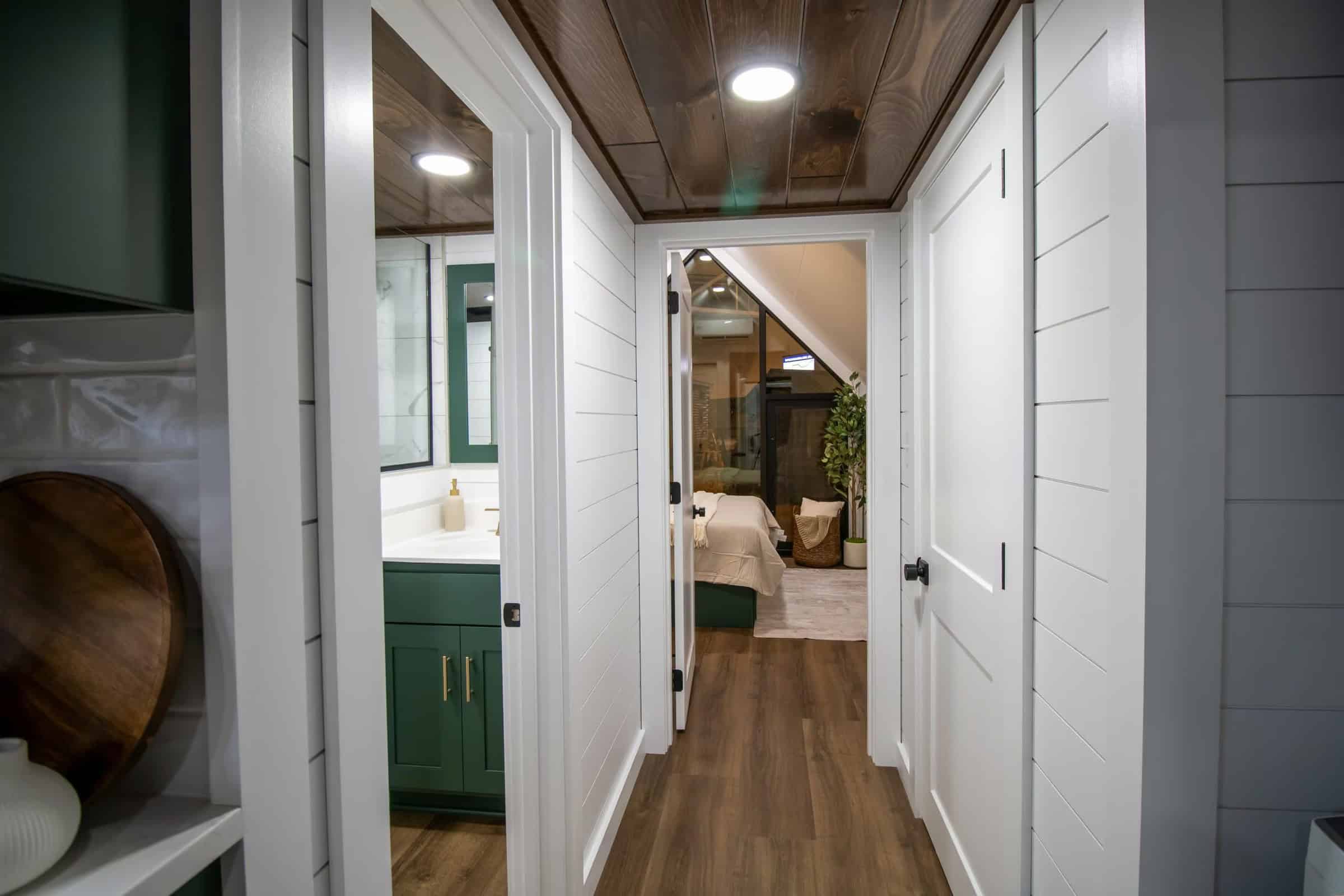 Hall toward main-floor bedroom: white shiplap, forest-green bath vanity glimpse, triangular A-frame bedroom window ahead