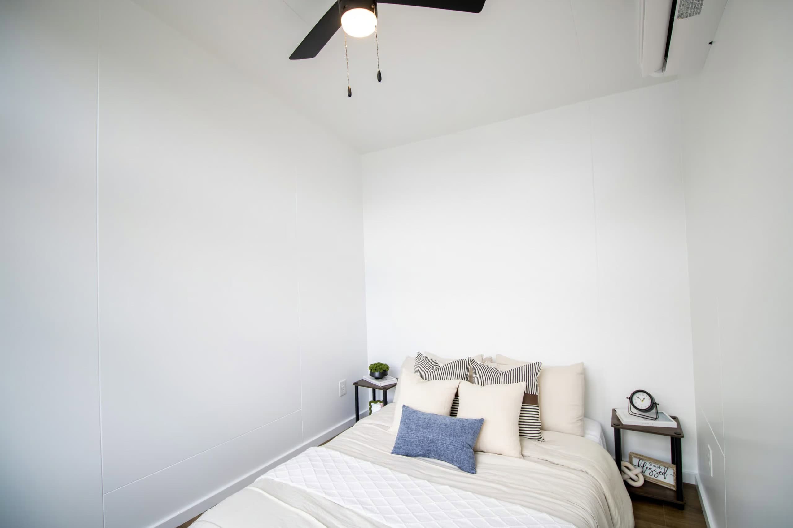 Main-floor bedroom: cream bedding, two dark side tables, black ceiling fan, white Bosch mini-split high on the painted-white wall