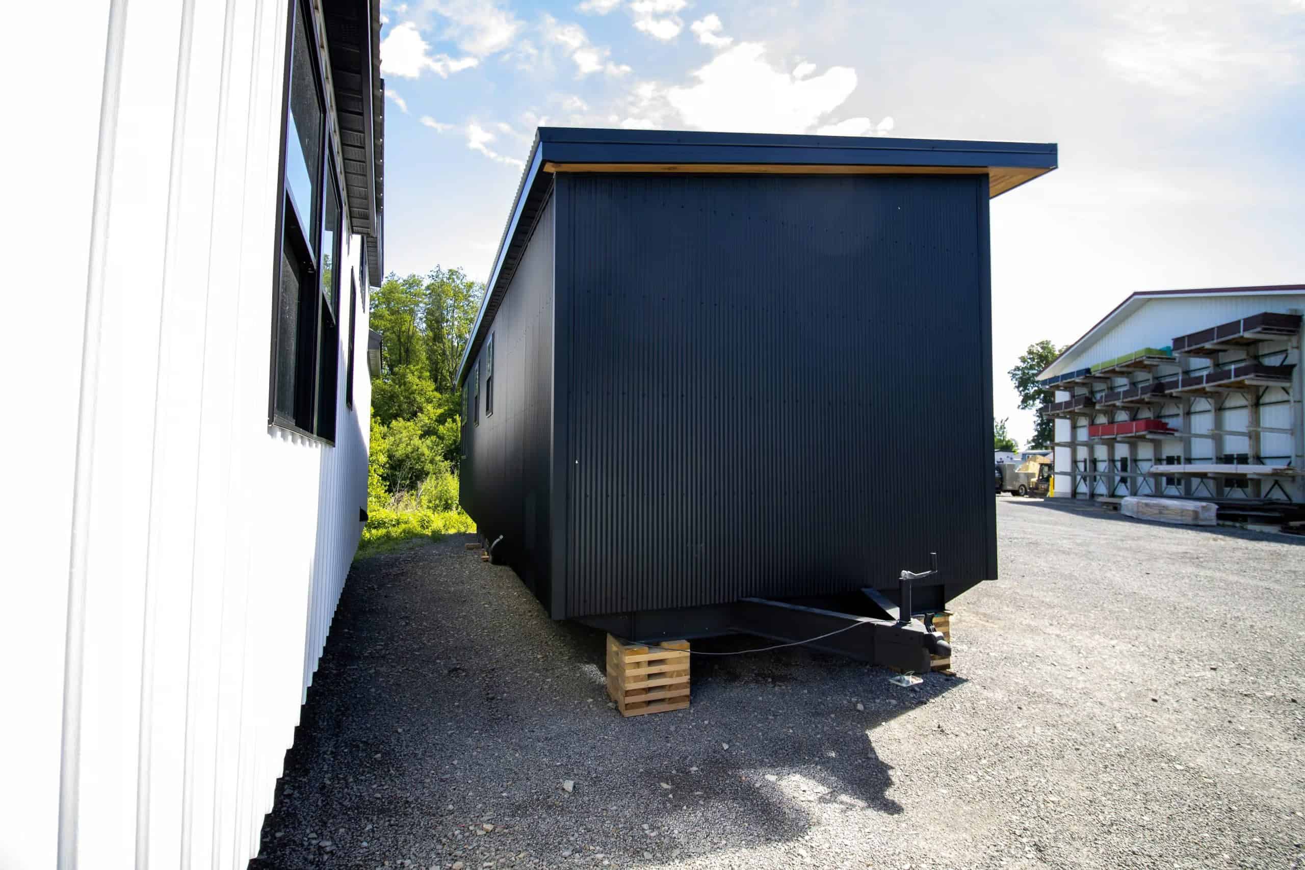 Rear three-quarter of the Modern Nova: black corrugated metal end wall, cedar soffit, steel trailer chassis on wood cribbing, hitch toward the shop building