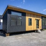 Modern Nova tiny house on a gravel lot: black corrugated metal, cedar around a full-glass entry door, black-framed windows, puck lights in the wood soffit