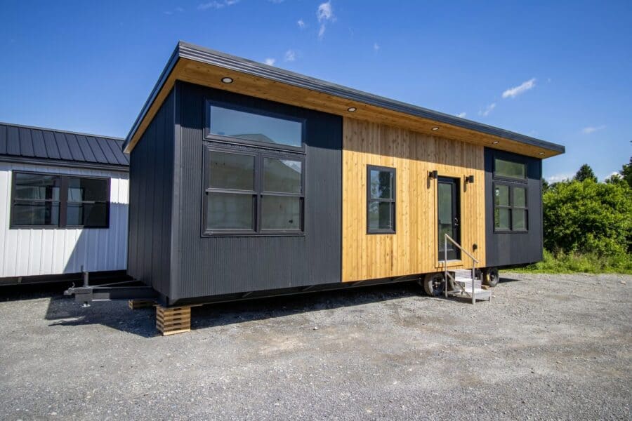 Modern Nova tiny house on a gravel lot: black corrugated metal, cedar around a full-glass entry door, black-framed windows, puck lights in the wood soffit